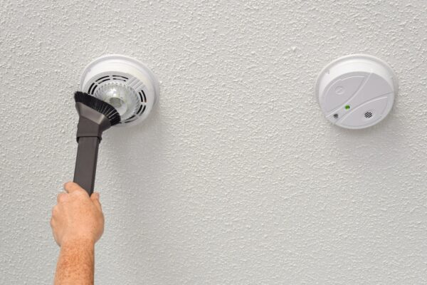 Person's hand vacuuming smoke and carbon monoxide detectors on a ceiling of a domestic room